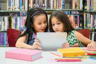 Two young girls read on a tablet in the library